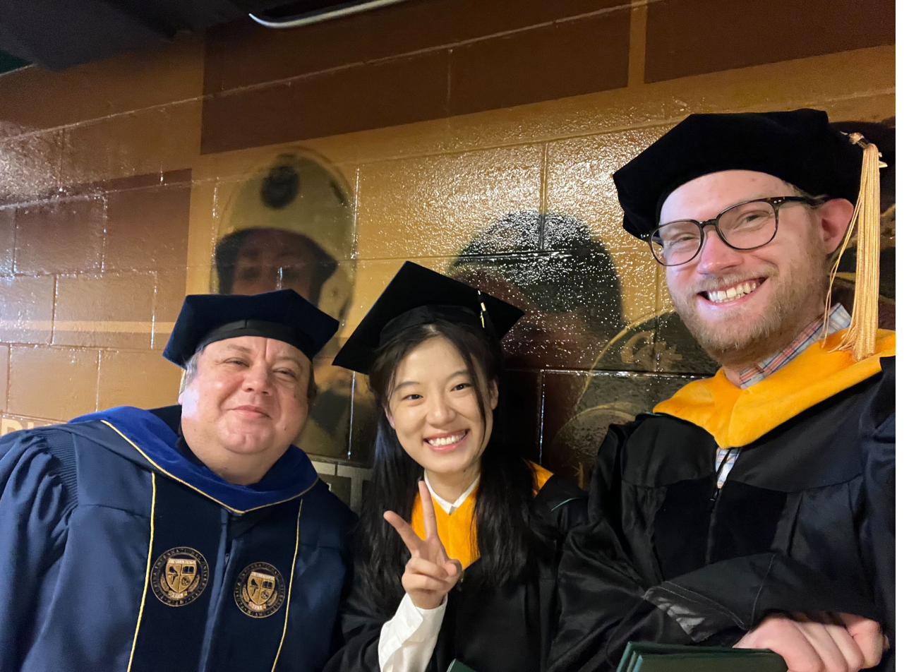 Francisco Ortega in doctoral regalia with two graduating students in caps and gowns at commencement, one flashing a peace sign.