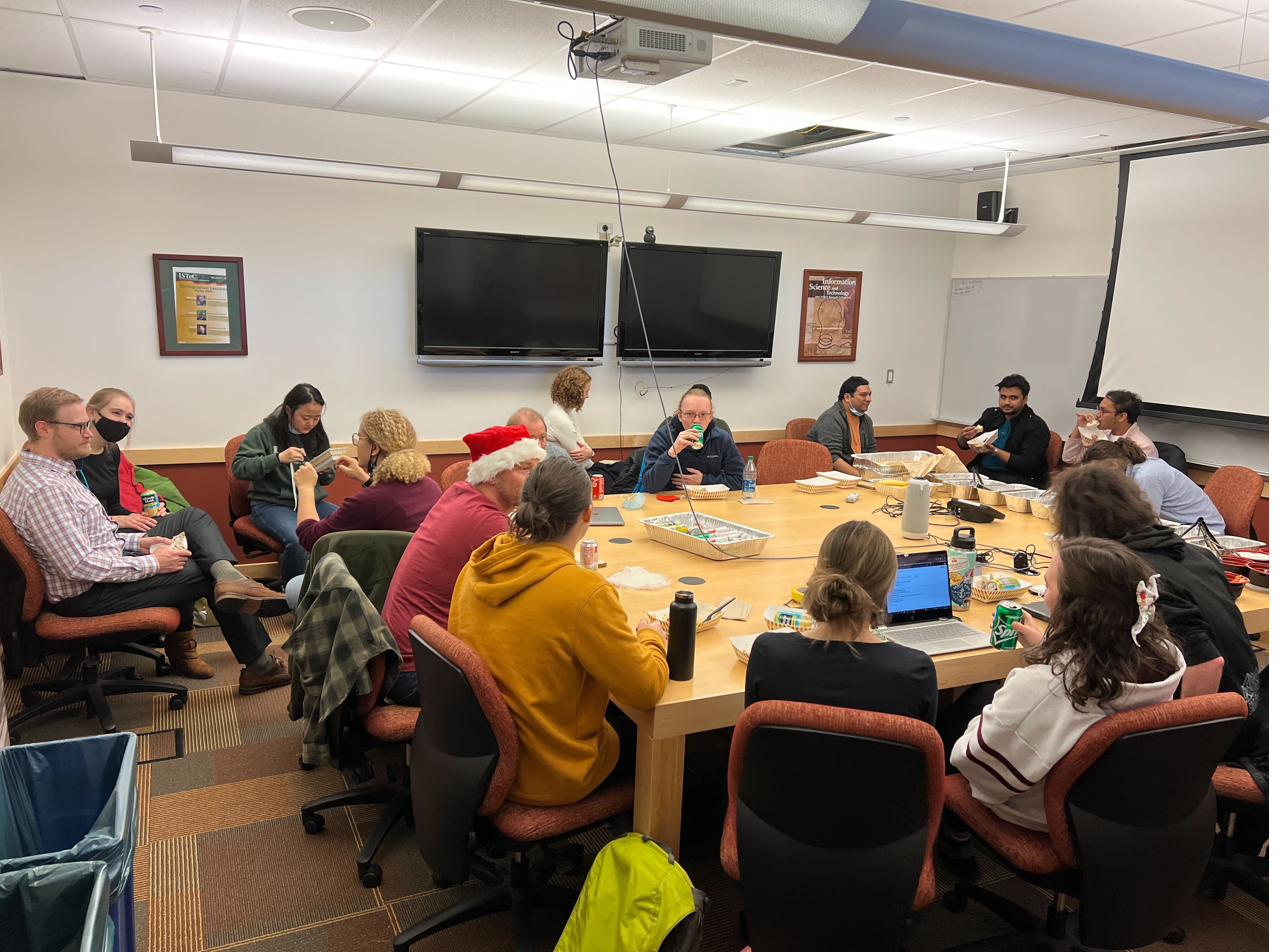 The research group around a large conference table sharing food during a holiday lab meeting, one member in a Santa hat.