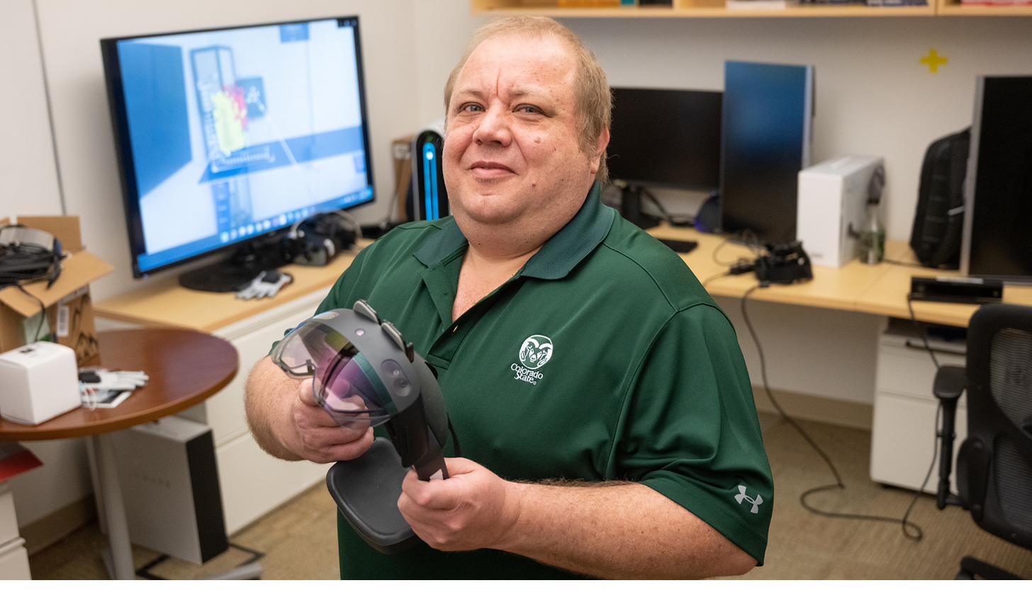 Francisco Ortega in a green Colorado State polo holding a HoloLens 2 headset in the lab, an AR scene visible on the monitor behind him.