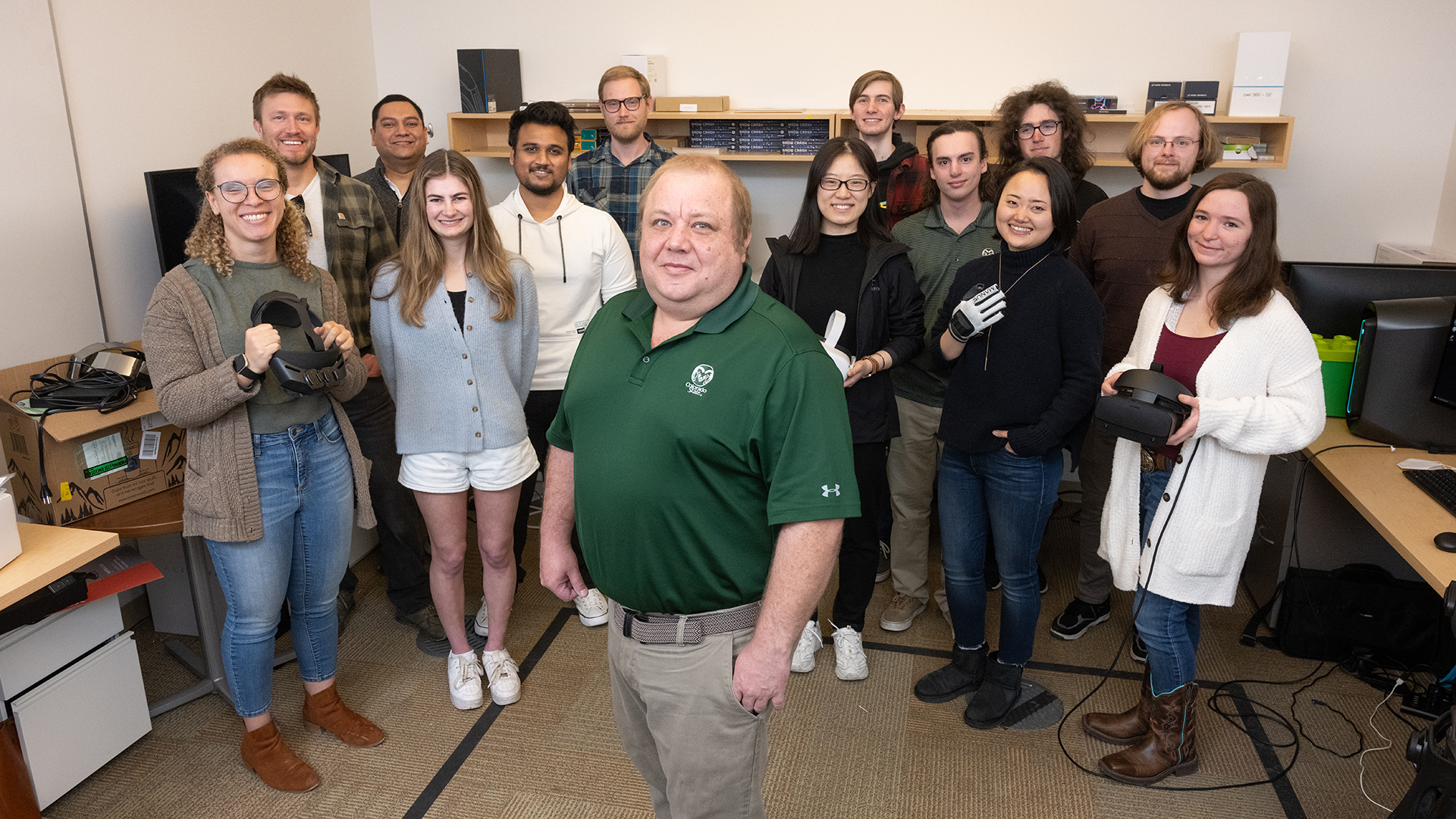 Fourteen lab members posing together in the lab around Francisco Ortega, several holding VR headsets and a haptic glove.