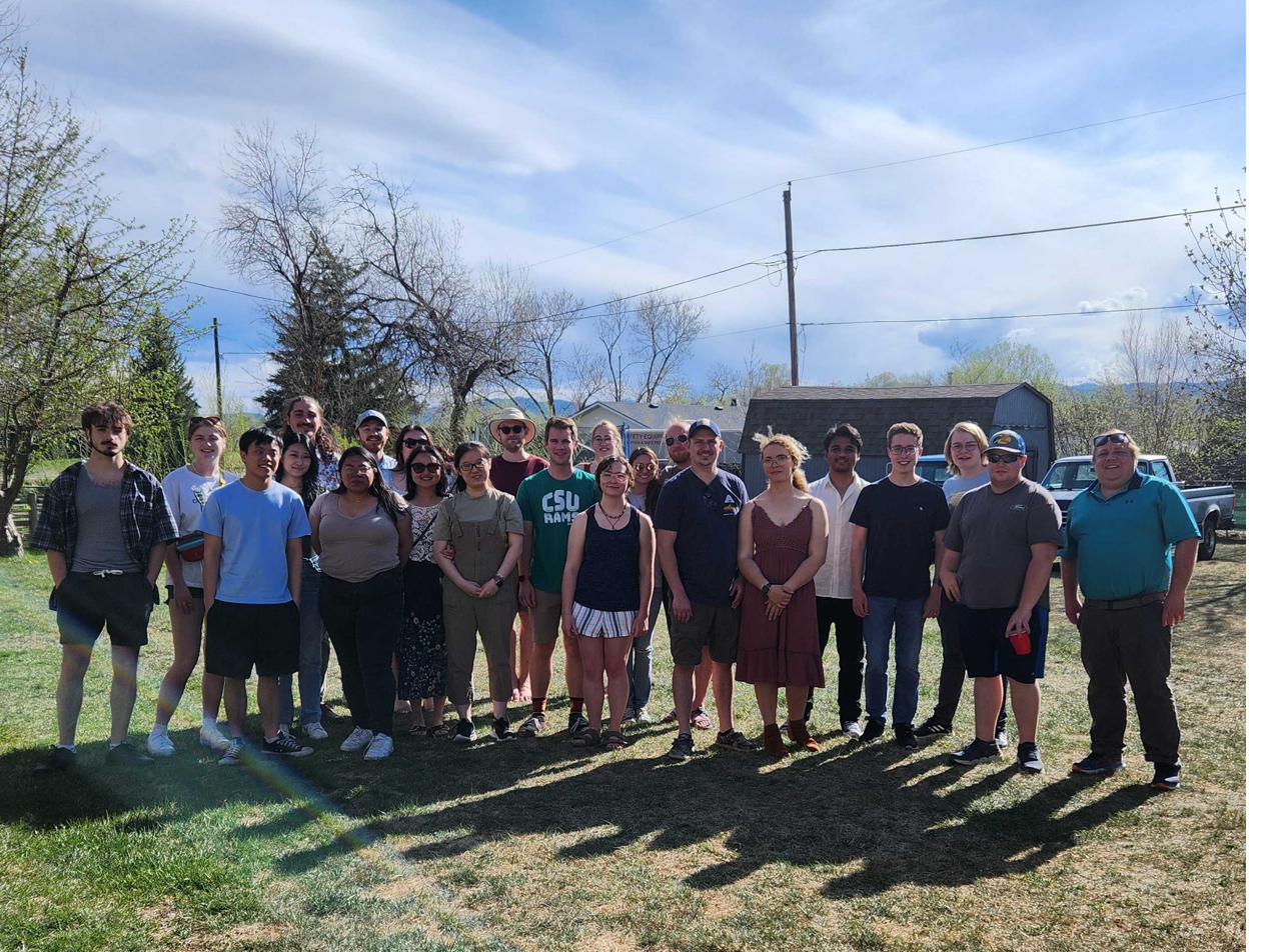 About twenty lab members and friends standing together on a sunny lawn at a spring get-together.